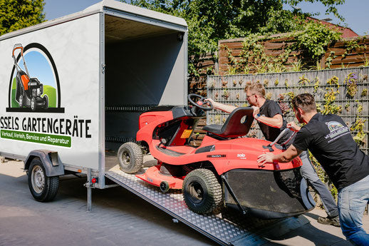 Zwei Personen laden einen roten Rasenmäher auf einen Lastwagen mit Rampe. Neben einem Anhänger mit Werbung "Wessels Gartengeräte" für Gartengeräte steht ein Anhänger.